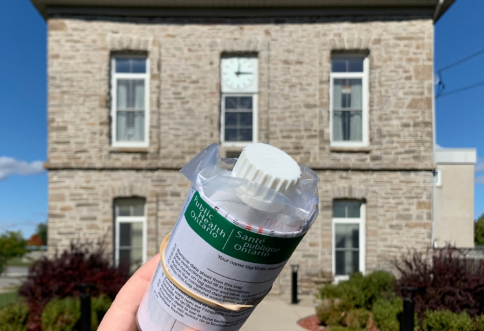 A hand holding onto a Public Health Ontario clear water sample jar in front of a stone building