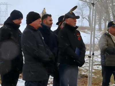 Several people stand in a row under a canopy during a snowy outdoor event with machinery and power lines in the background.