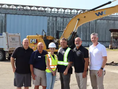 Six individuals, including municipal leaders and project representatives, pose in front of a WK excavator and CAT equipment at the Port of Johnstown.