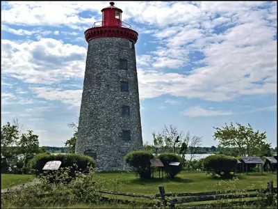 Stone lighthouse with a red top surrounded by signs and greenery by water.