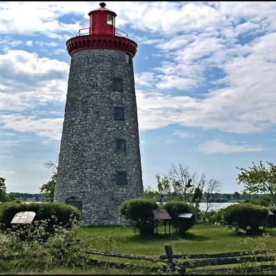 Stone lighthouse with a red top surrounded by signs and greenery by water.