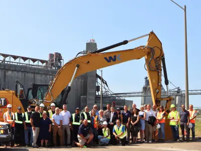Large group of workers, partners, and officials standing in front of a WK excavator at the Port of Johnstown. The industrial grain terminal structure is visible in the background.