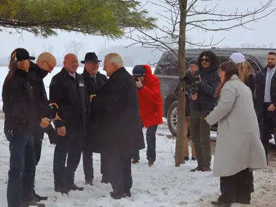People stand outdoors on snowy ground talking near parked vehicles while photographers and staff observe.