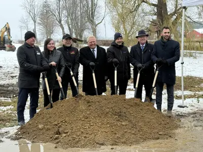 A group of seven people stand outdoors at a groundbreaking event. They are holding shovels in front of a mound of soil with light snow on the ground and trees in the background.