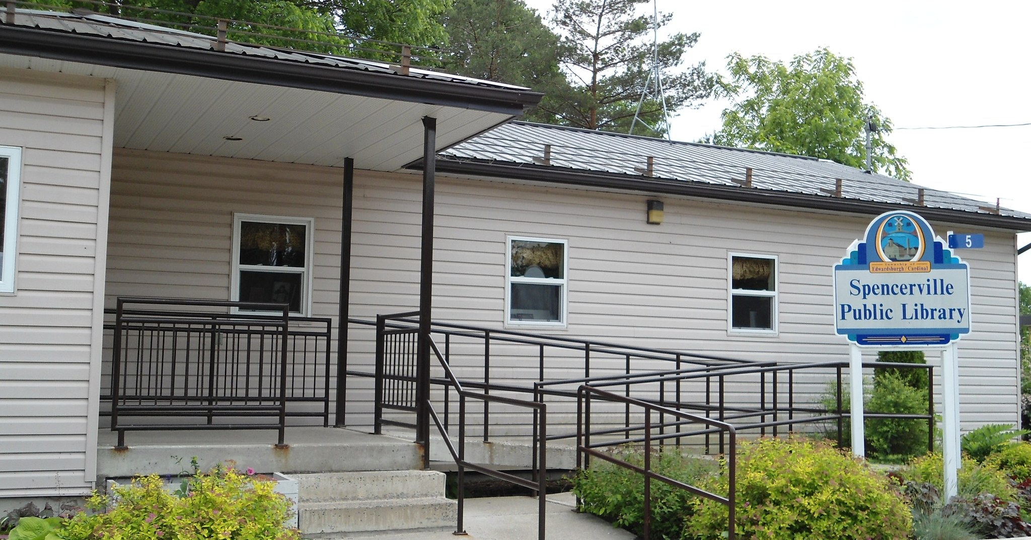 Exterior view of the Spencerville Branch of the Edwardsburgh Cardinal Public Library showing the main entrance and accessibility ramp with metal railings and surrounding greenery.