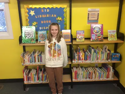 A girl in a Lion King hoodie stands smiling in front of a library book display titled “Kid Librarian Book Display,” surrounded by colorful children’s books.