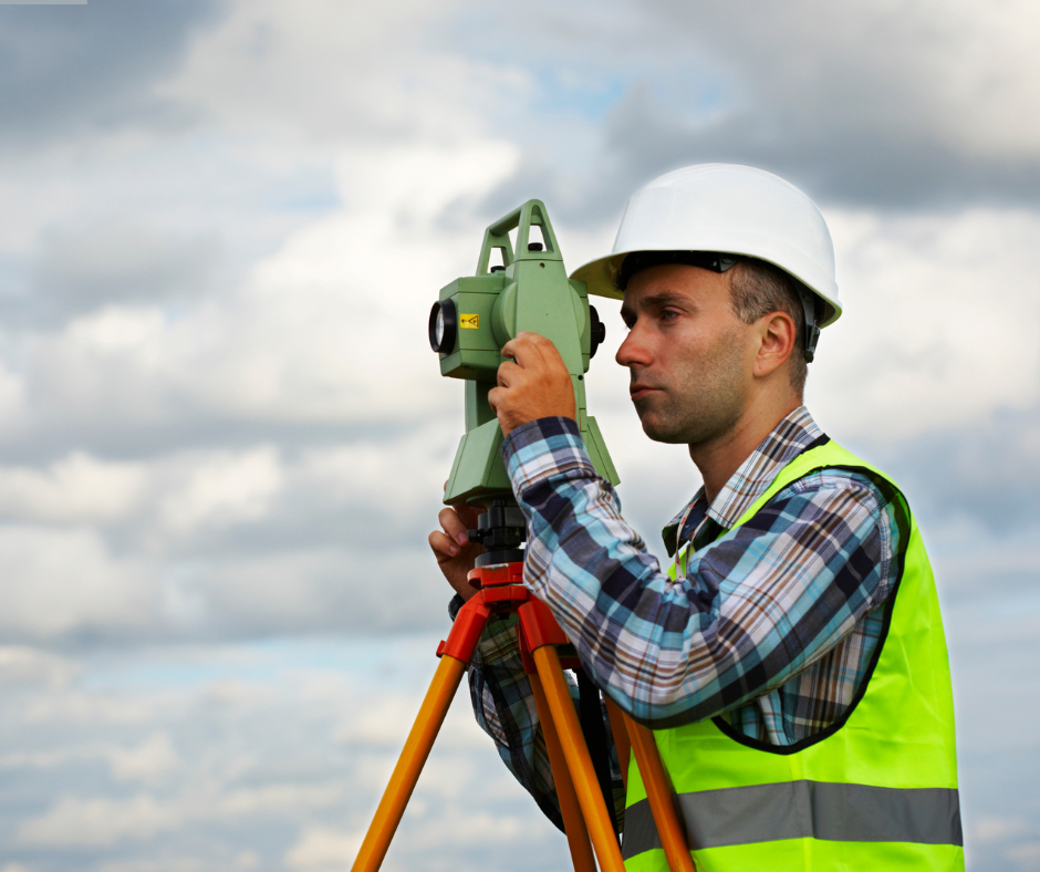 Man in safety vest and hard hat using a theodolite on a tripod outdoors.