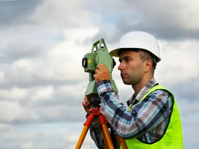 Man in safety vest and hard hat using a theodolite on a tripod outdoors.