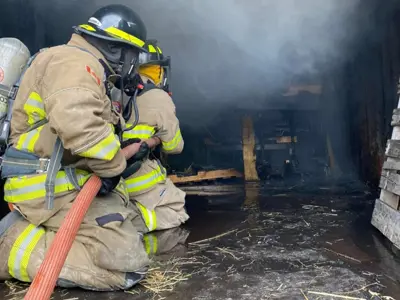 Two firefighters in full protective gear and helmets kneel on a wet floor, aiming a fire hose into a smoke-filled training area with wooden pallets and charred debris.