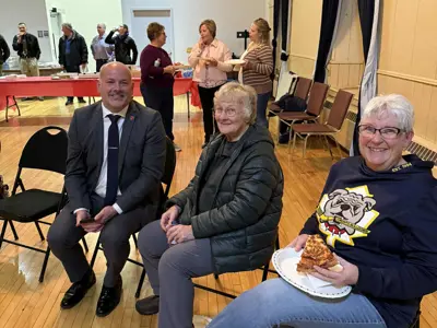 People seated at an event talking and eating while others gather behind.