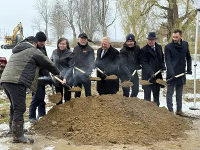 A group of people stand outdoors in winter conditions holding shovels of soil during a groundbreaking event, with smoke rising from an Indigenous smudging ceremony taking place in front of them.