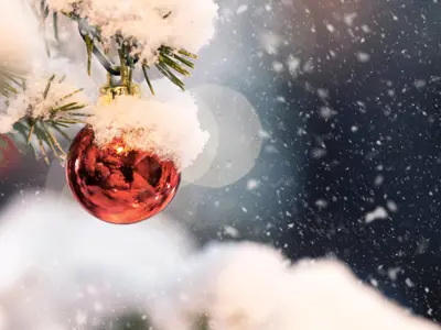 A close-up of a red Christmas ornament hanging from a snow-covered evergreen branch. Soft snowflakes fall against a blurred winter background, creating a peaceful and festive holiday atmosphere.