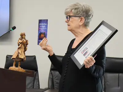 Woman at a podium holding a book and framed write-up, with a sculpture nearby.