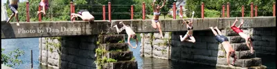 Group of youth jumping off an old stone bridge into a river on a sunny day.