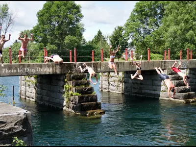 Group of youth jumping off an old stone bridge into a river on a sunny day.