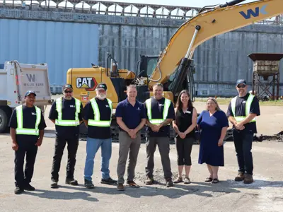 V6 Agronomy and project team members stand in front of excavation equipment and a WK truck at the Port of Johnstown. A concrete grain terminal wall is in the background.